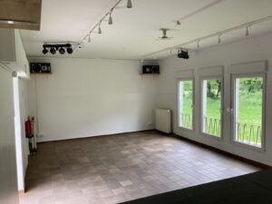 Vereinsheim Hauptraum mit braunen Bodenfliesen und Fenstern. Weiße Wände, Deckenbeleuchtungn.